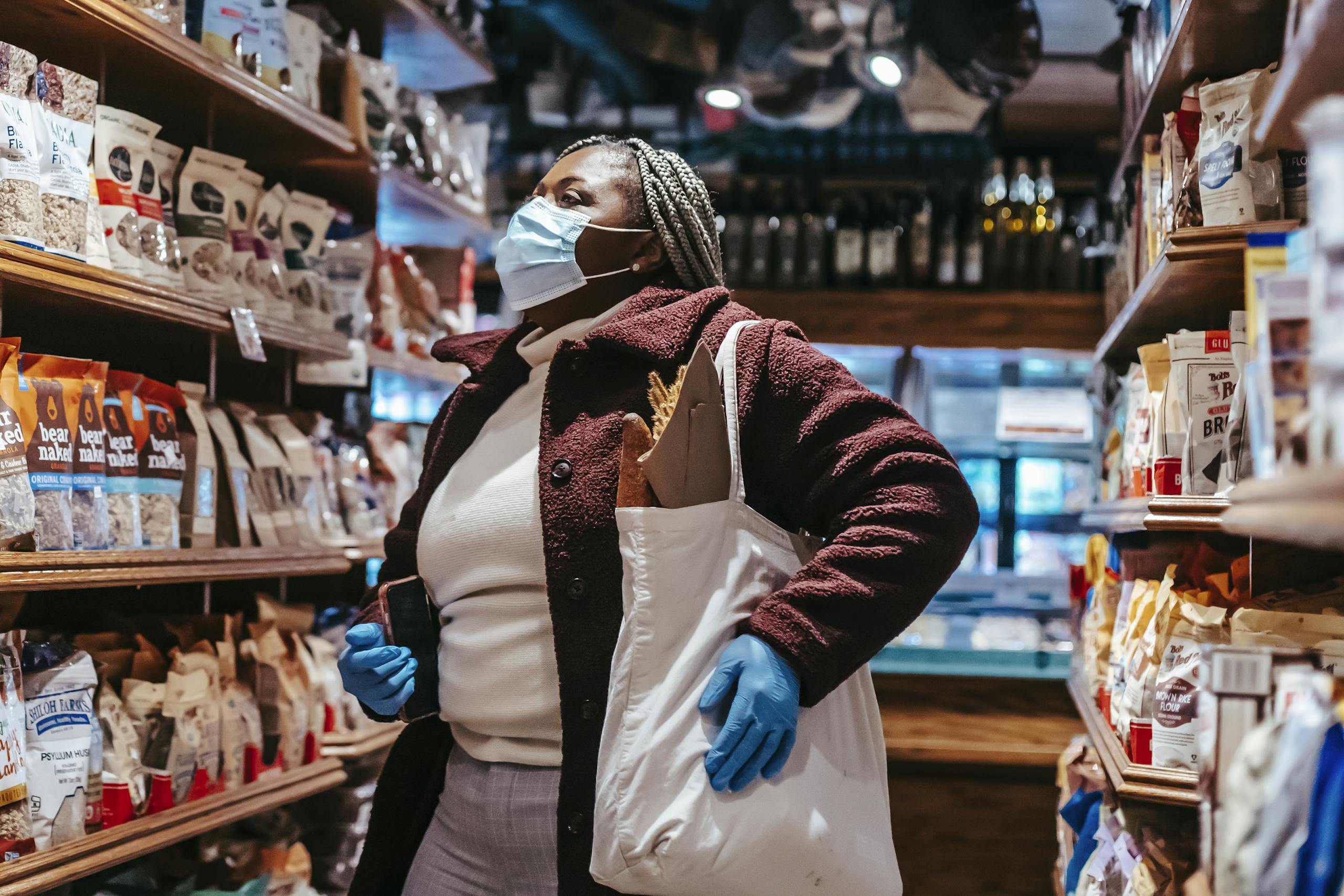 African American female buyer with shopping bag and smartphone in protective mask and gloves selecting products in supermarket